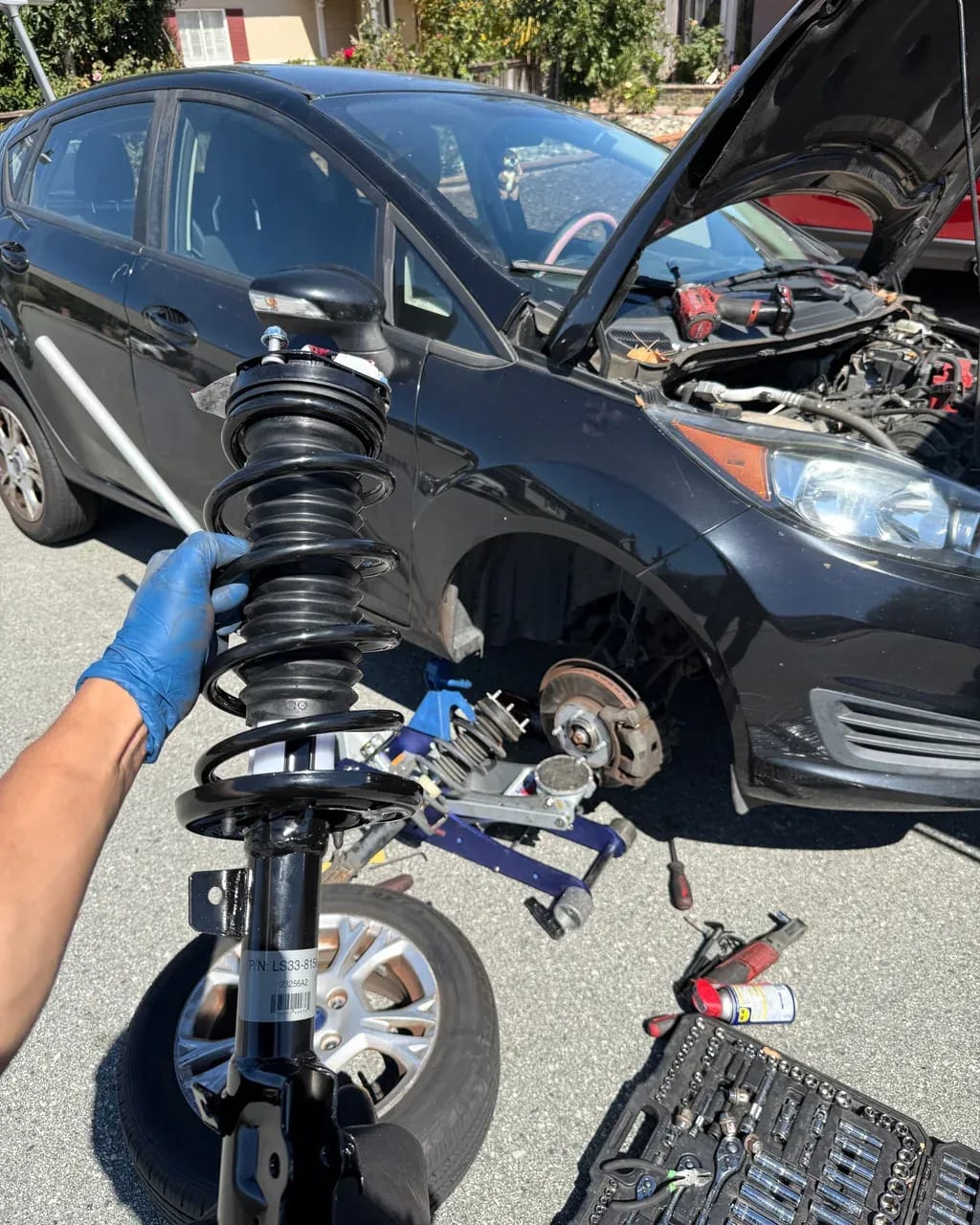 Stewart mechanic holding a new strut assembly next to a Ford Fiesta during a mobile suspension repair