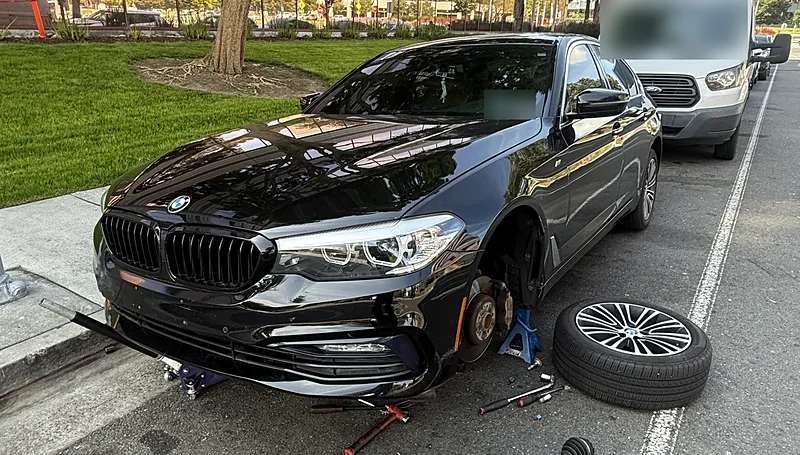 BMW 5 Series on a jack stand with the front wheel off during a mobile front-end repair outside an office