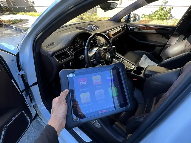 Autel scan tablet held at the open driver's door of a white Porsche Macan during mobile check engine light diagnostics