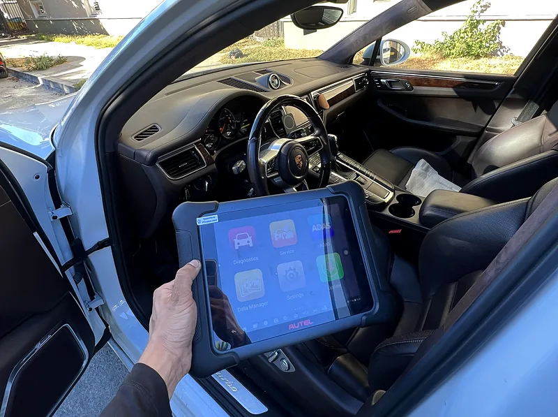 Autel scan tablet held at the open driver's door of a white Porsche Macan during mobile check engine light diagnostics